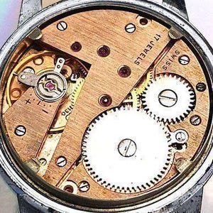 Op de afbeelding: Close-up van een uurwerk met 17 juwelen en de tekst "Swiss" gedrukt op de plaat. Het uurwerk is gemaakt van messing en heeft een gouden tint. Het uurwerk is zichtbaar door de achterkant van de horlogekast.
