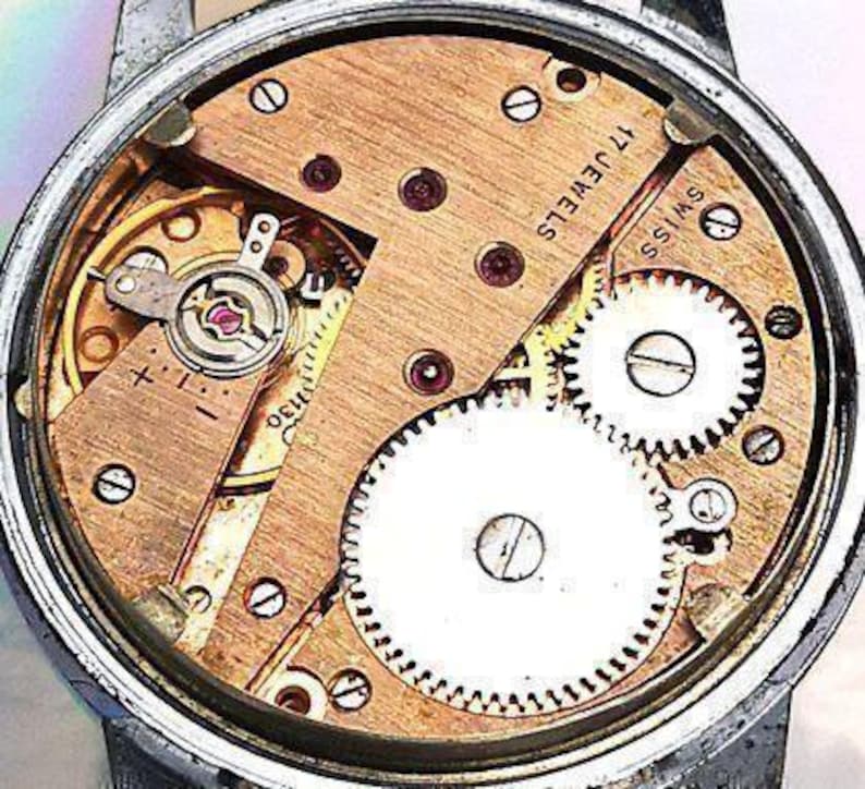 Op de afbeelding: Close-up van een uurwerk met 17 juwelen en de tekst "Swiss" gedrukt op de plaat. Het uurwerk is gemaakt van messing en heeft een gouden tint. Het uurwerk is zichtbaar door de achterkant van de horlogekast.
