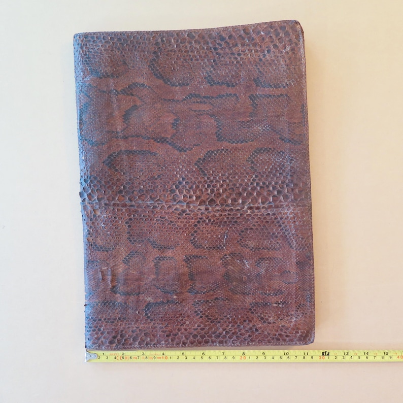 K&ouml;nnte beinhalten: Eine braune Leder-Notizbuchh&uuml;lle mit Schlangenmuster. Die H&uuml;lle ist rechteckig und etwa 25 cm lang. Die H&uuml;lle hat eine strukturierte Oberfl&auml;che und ein dunkelbraunes und schwarzes Muster.