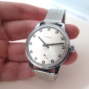 Op de afbeelding: Een zilveren polshorloge met een witte wijzerplaat en zwarte wijzers. Het horloge heeft een zilveren metalen band en op de wijzerplaat staat "Hoffmann 17 Rubis Incabloc".