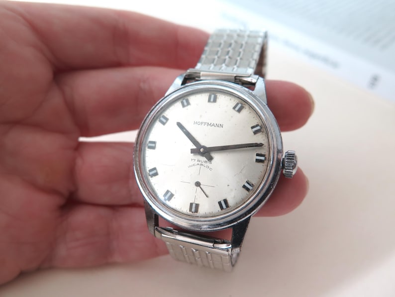 Op de afbeelding: Een zilveren polshorloge met een witte wijzerplaat en zwarte wijzers. Het horloge heeft een zilveren metalen band en op de wijzerplaat staat "Hoffmann 17 Rubis Incabloc".