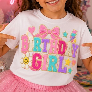 May include: A white short-sleeved t-shirt featuring a colorful graphic print that reads "BIRTHDAY GIRL" in large, textured, glittery letters. The design includes a pink bow, a yellow smiley face flower, stars, and a lightning bolt.