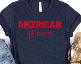 May include: Navy blue t-shirt with the words "AMERICAN Woman" in red. The shirt is styled with denim shorts, sunglasses, and gold sandals. The outfit is laid out on a white background.