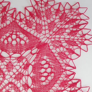 May include: A close-up of a pink crocheted doily with a delicate, intricate pattern. The doily is made with fine yarn and has a scalloped edge.
