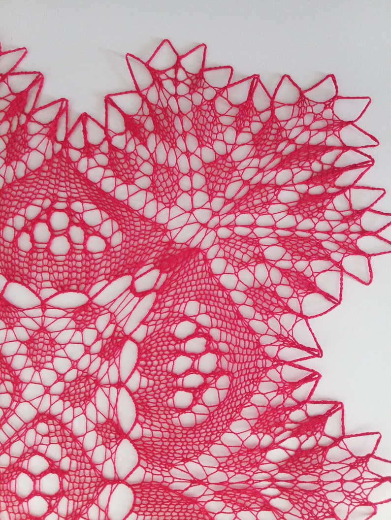 May include: A close-up of a pink crocheted doily with a delicate, intricate pattern. The doily is made with fine yarn and has a scalloped edge.
