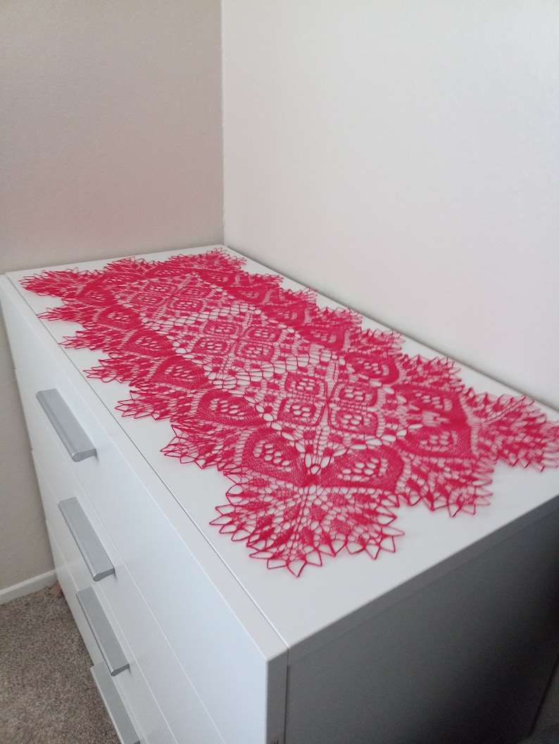 May include: A pink crocheted table runner with a delicate lace pattern. The runner is laid out on a white dresser with three drawers.