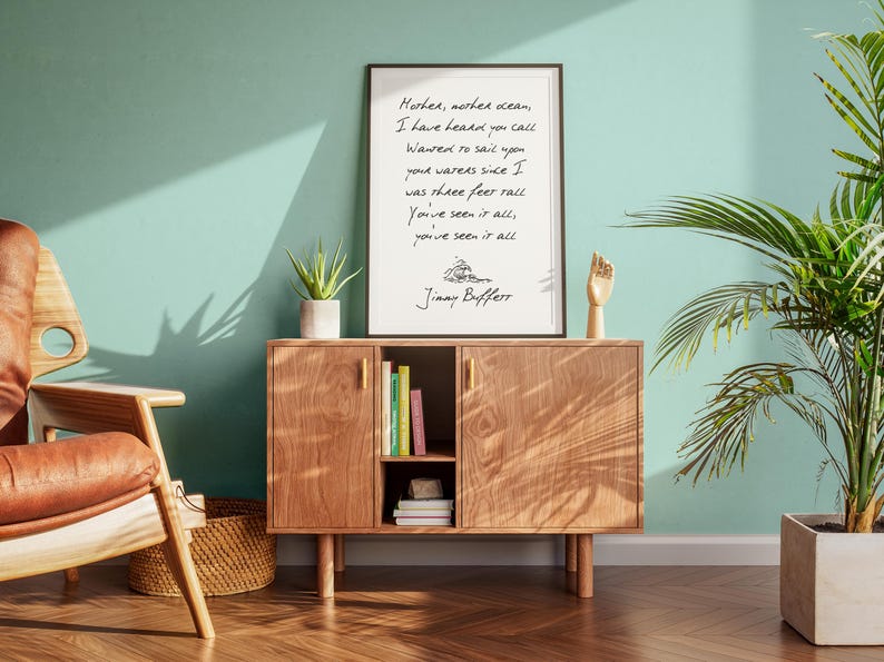 May include: A framed print with a handwritten quote by Jimmy Buffett, "Mother, mother ocean, I have heard you call, wanted to sail upon your waters since I was three feet tall. You've seen it all, you've seen it all." The print is on a wooden shelf with books and a plant.