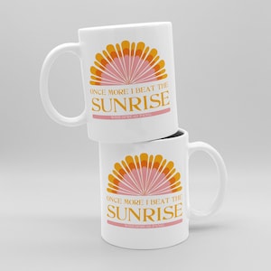 May include: Two white ceramic mugs stacked on top of each other. Each mug features a sunrise design in orange and pink, with the text "ONCE MORE I BEAT THE SUNRISE" in orange and pink.
