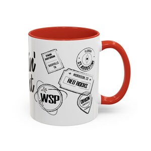 May include: White ceramic mug with a red rim and handle. The mug features a black and white design of various travel stamps and the text "It's in It".