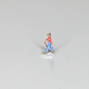 May include: A miniature figurine of a person wearing a red and white striped shirt, blue jeans, and a red hat. The figurine is holding a stick and is standing on a small white base.