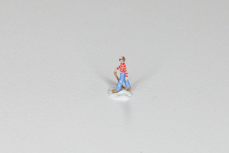 May include: A miniature figurine of a person wearing a red and white striped shirt, blue jeans, and a red hat. The figurine is holding a stick and is standing on a small white base.