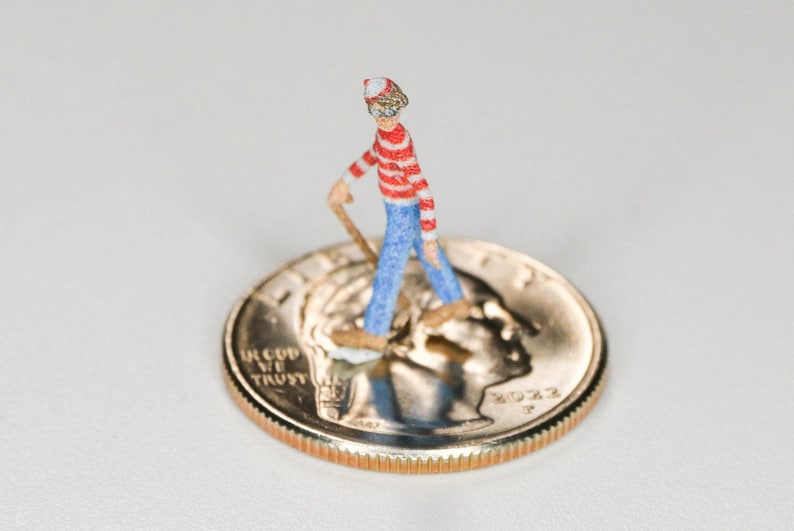 May include: A miniature figurine of a person wearing a red and white striped shirt, blue jeans, and a red and white hat, standing on a US quarter with the year 2022.
