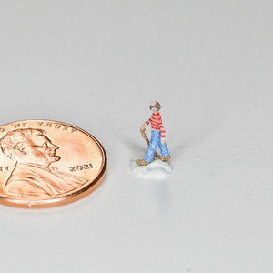 May include: A miniature figurine of a person wearing a red and white striped shirt, blue jeans, and a red hat. The figurine is holding a stick and standing on a small white base. A 2021 US penny is in the background for scale.