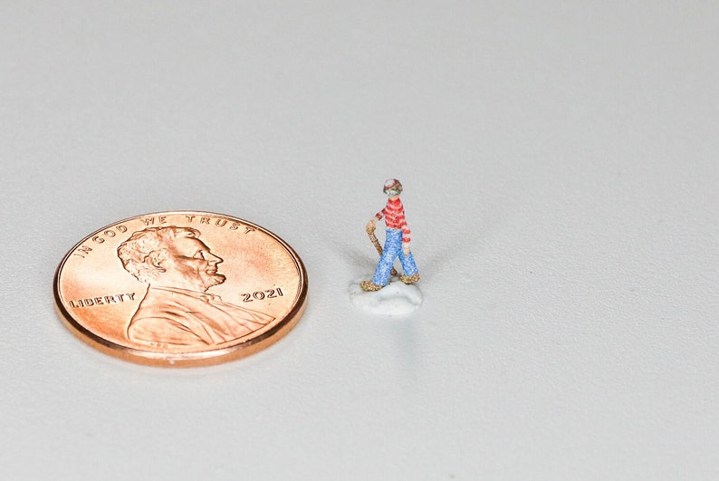 May include: A miniature figurine of a person wearing a red and white striped shirt, blue jeans, and a red hat. The figurine is holding a stick and standing on a small white base. A 2021 US penny is in the background for scale.