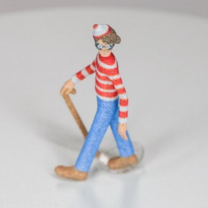 May include: A small, red and white striped figurine of a person wearing a red hat and glasses, holding a wooden cane. The figurine is walking forward.