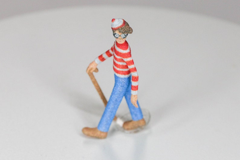 May include: A small, red and white striped figurine of a person wearing a red hat and glasses, holding a wooden cane. The figurine is walking forward.