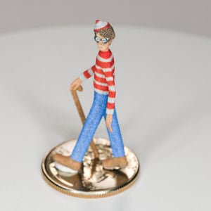 May include: A small, red and white striped figurine of a person wearing a red hat and glasses, holding a walking stick and standing on a gold coin.