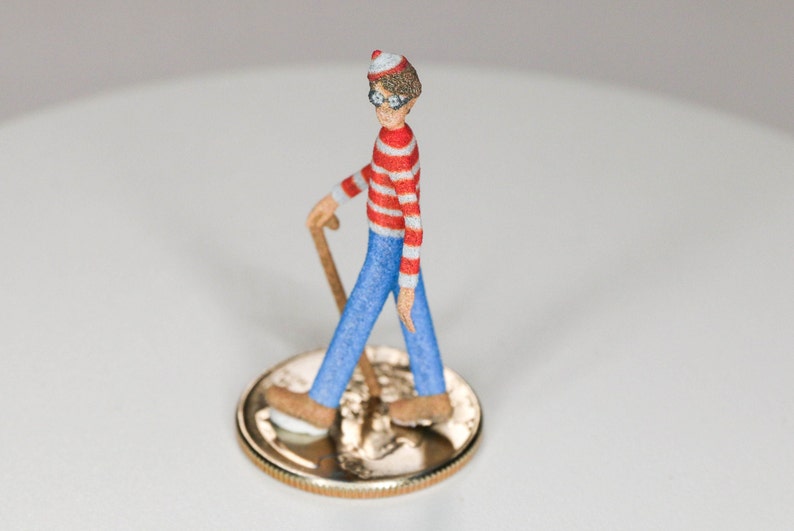 May include: A small, red and white striped figurine of a person wearing a red hat and glasses, holding a walking stick and standing on a gold coin.