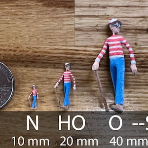 May include: Three miniature figures of a person wearing a red and white striped shirt and blue pants, holding a stick. The figures are in different sizes, measuring 10 mm, 20 mm, and 40 mm. The figures are on a wooden surface.