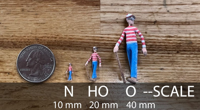 May include: Three miniature figures of a person wearing a red and white striped shirt and blue pants, holding a stick. The figures are in different sizes, measuring 10 mm, 20 mm, and 40 mm. The figures are on a wooden surface.