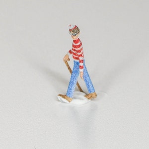 May include: A small figurine of a person wearing a red and white striped shirt, blue jeans, and a red and white hat. The person is holding a stick and is walking on a white surface.