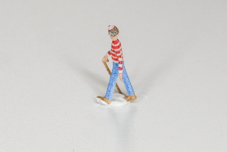 May include: A small figurine of a person wearing a red and white striped shirt, blue jeans, and a red and white hat. The person is holding a stick and is walking on a white surface.