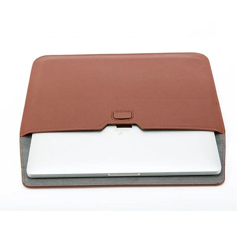 May include: A brown leather laptop sleeve with a gray felt lining. The sleeve is closed and a laptop is visible inside.