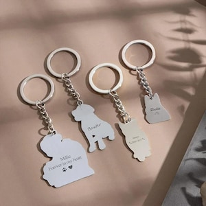 May include: Silver keychains with dog and cat silhouettes. Each keychain has a chain and a ring. Some keychains have engraved names and dates, and one says "Millie Forever in my heart" with paw prints and a heart.