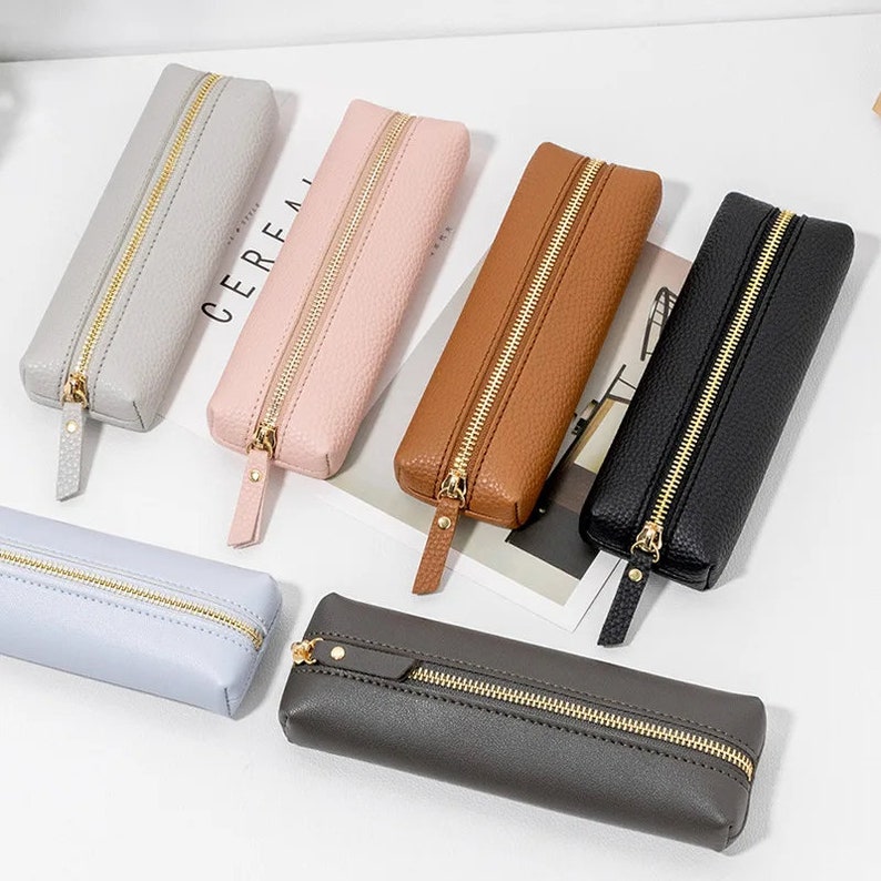 May include: Six zippered pencil cases in various colors, including gray, pink, brown, black, and light blue. The cases are made of a textured material and have gold zippers.