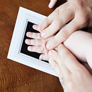 May include: A white framed ink pad with a black surface is used to capture a baby's handprint. The baby's hand is pressed onto the ink pad, guided by adult hands. The wooden surface provides a warm contrast.