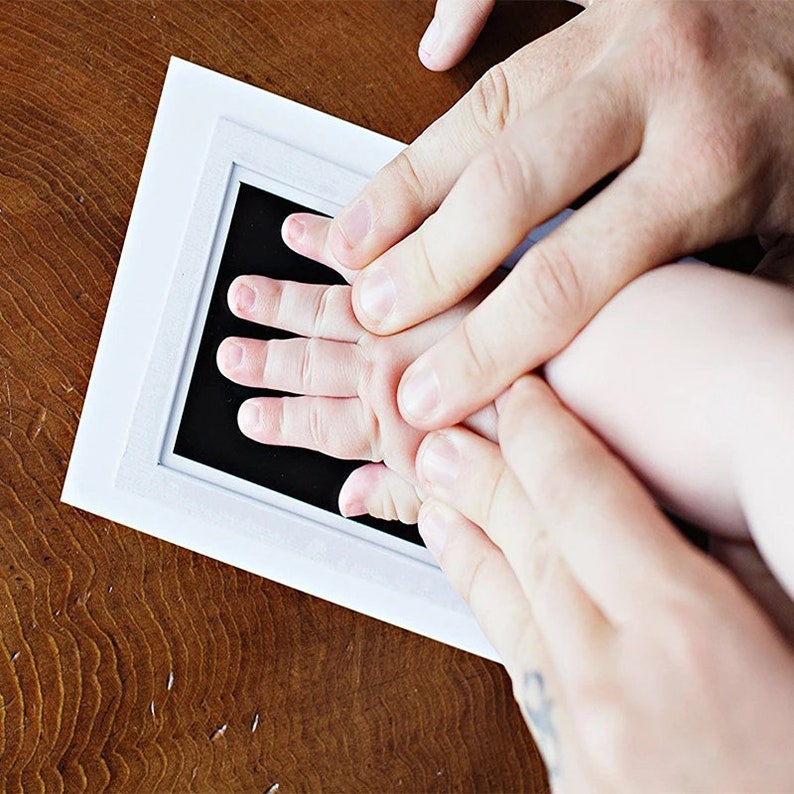 May include: A white framed ink pad with a black surface is used to capture a baby's handprint. The baby's hand is pressed onto the ink pad, guided by adult hands. The wooden surface provides a warm contrast.