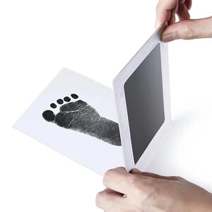 May include: A baby footprint kit with a black ink pad and a white card. The card displays a detailed black footprint. The ink pad is being lifted to reveal the print. The kit is designed for creating baby keepsakes.