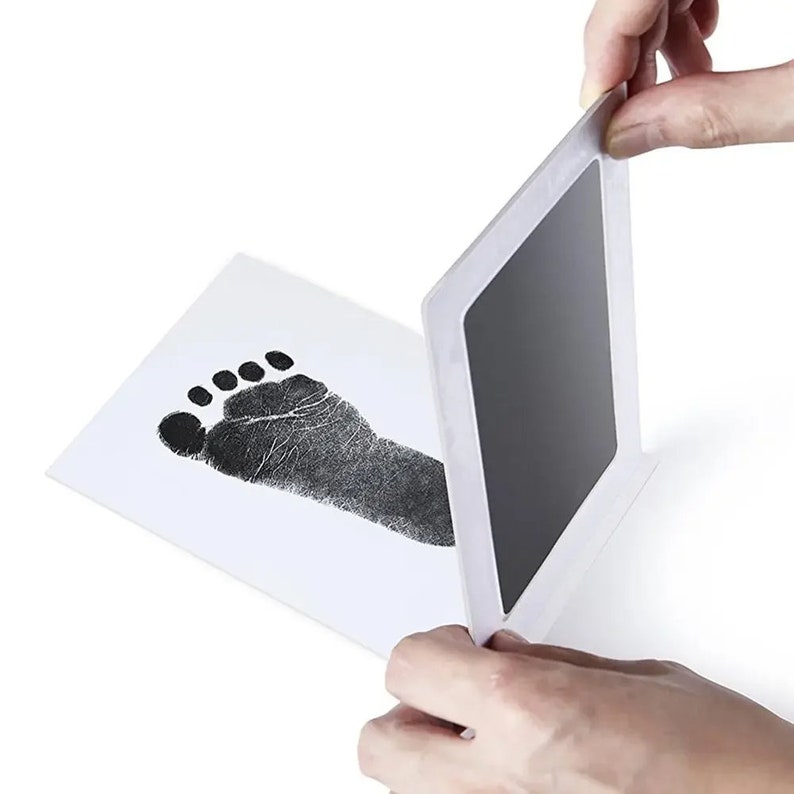 May include: A baby footprint kit with a black ink pad and a white card. The card displays a detailed black footprint. The ink pad is being lifted to reveal the print. The kit is designed for creating baby keepsakes.