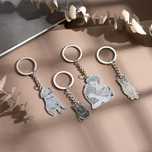 May include: Four silver keychains with detailed engraved pendants. The pendants feature a dog, a person hugging a dog, a dog, and a cat. Each keychain has a silver ring and a chain.