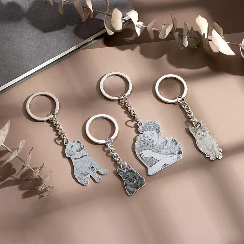 May include: Four silver keychains with detailed engraved pendants. The pendants feature a dog, a person hugging a dog, a dog, and a cat. Each keychain has a silver ring and a chain.