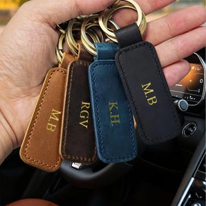 May include: Four rectangular leather keychains in tan, dark brown, blue, and black, each featuring gold-stamped initials. The tan keychain is marked M.B, the dark brown one RGV, the blue one K.H, and the black one M.B. Each has a gold metal ring.