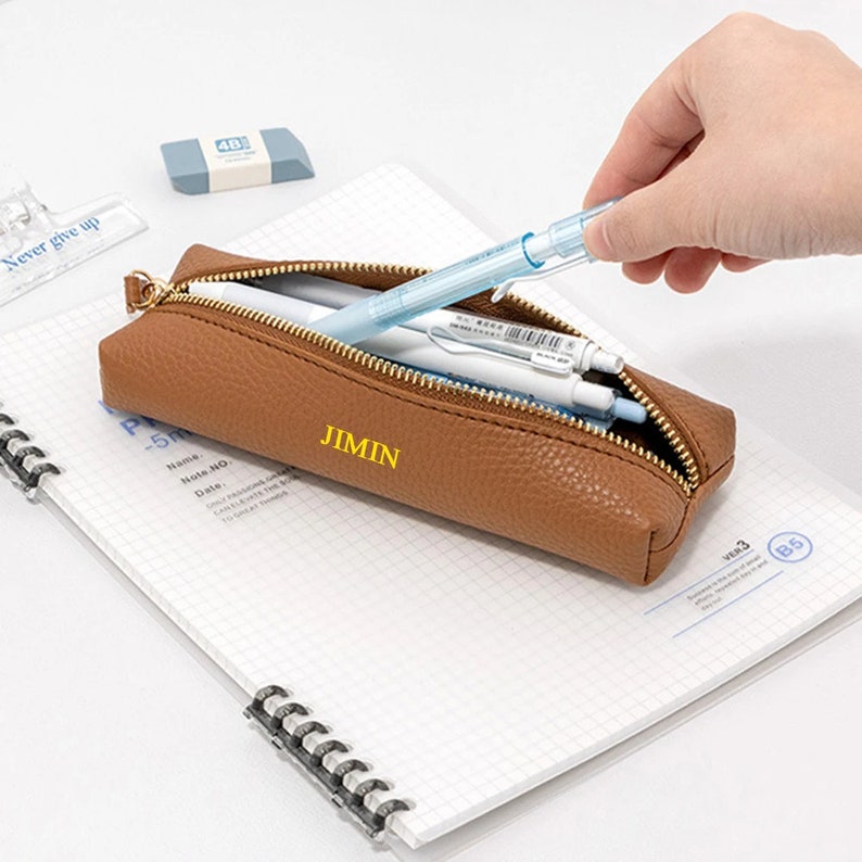 May include: A brown leather pencil case with a gold zipper. The case is open and has several pens and pencils inside. The case has the name "JIMIN" printed on it.