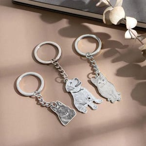 May include: Three silver keychains with detailed pet portraits: a German Shepherd, a Labrador, and a cat. Each keychain features a circular ring and a chain connecting to the pet-shaped pendant. The keychains are displayed on a neutral-toned surface.