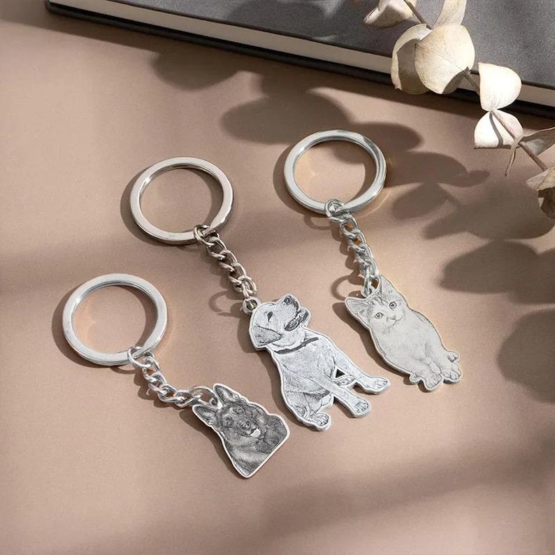 May include: Three silver keychains with detailed pet portraits: a German Shepherd, a Labrador, and a cat. Each keychain features a circular ring and a chain connecting to the pet-shaped pendant. The keychains are displayed on a neutral-toned surface.