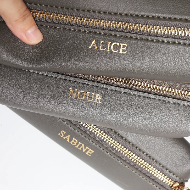 May include: Three gray zippered pouches with gold zippers. The pouches are stacked on top of each other. The top pouch has the name "ALICE" written in gold lettering. The middle pouch has the name "NOUR" written in gold lettering. The bottom pouch has the name "SABINE" written in gold lettering.