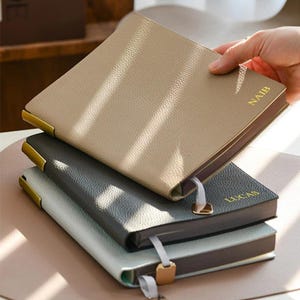 Op de afbeelding: Een stapel van drie rechthoekige notitieboeken met gestructureerde covers in beige, donkergrijs en lichtgrijs. Elk notitieboek heeft goudkleurige metalen hoeken, een lintbladwijzer met een gouden bedel en de goudkleurige namen NAIB en LUCAS.