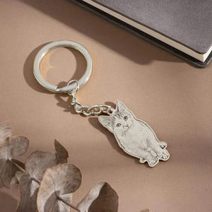May include: Silver-colored cat-shaped keychain. The keychain features a detailed engraving of a cat with a round keyring and a chain. The cat is lying down with its paws extended. The background is a neutral color.