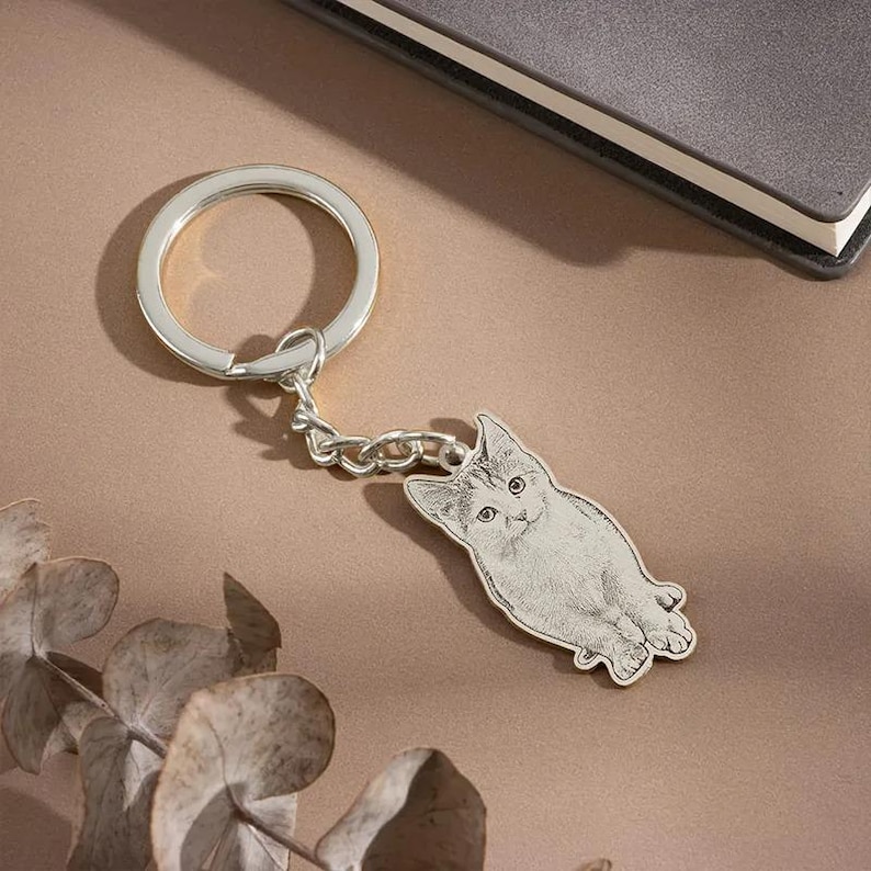 May include: Silver-colored cat-shaped keychain. The keychain features a detailed engraving of a cat with a round keyring and a chain. The cat is lying down with its paws extended. The background is a neutral color.