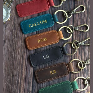 May include: A collection of rectangular leather keychains in red, teal, tan, black, brown, and green, each featuring a metal split ring and a lobster clasp. The teal keychain is embossed with CALLUM, tan with ROB, black with LG, and brown with S.R.