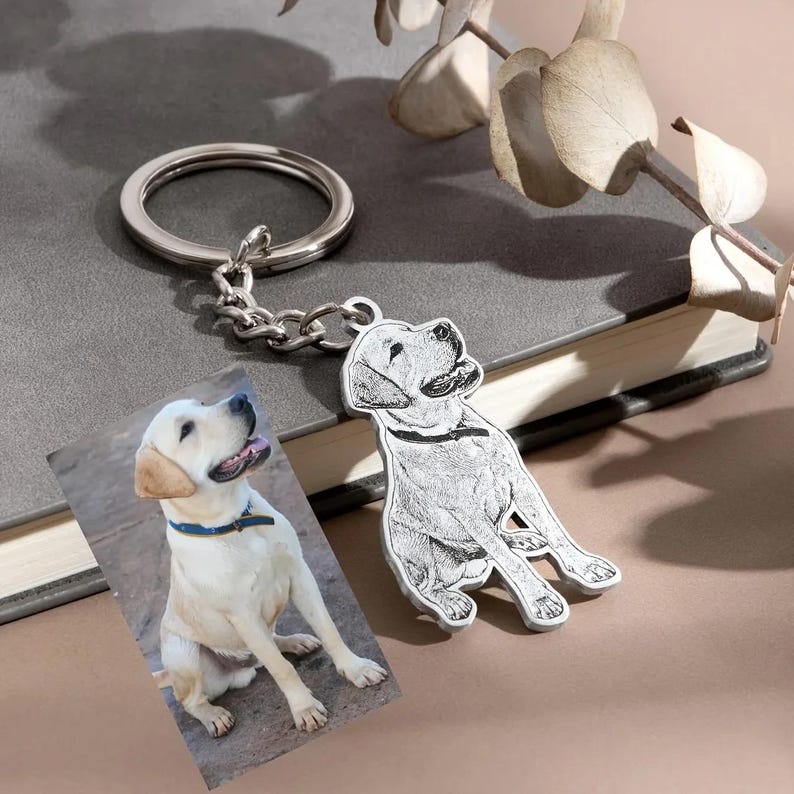 May include: A silver-colored keychain with a dog pendant and a photo of a Labrador Retriever. The pendant is a detailed sketch of a dog. The photo shows a yellow Labrador Retriever wearing a blue collar.