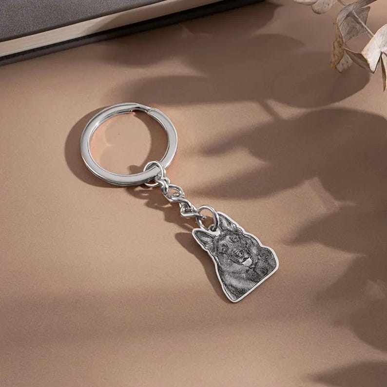 May include: Silver-colored keychain featuring a detailed pendant of a German Shepherd dog. The pendant is attached to a chain and a large key ring. The dog's image is in grayscale, with visible fur and facial features.