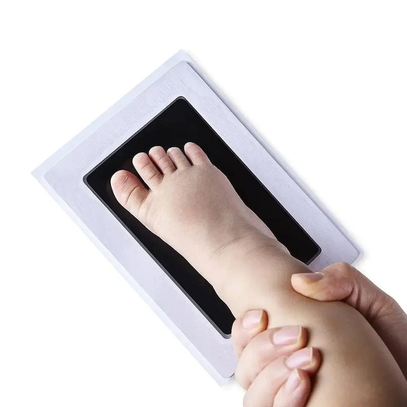 May include: A baby's foot being pressed onto a black ink pad within a white frame. The pad is designed to capture a footprint. The image is taken from a slightly elevated angle, with the baby's foot and ankle visible.
