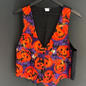 May include: A Halloween-themed vest with a black back and a vibrant front featuring orange jack-o'-lanterns on a purple background. The vest has a V-neck and is buttoned up, perfect for a festive outfit.