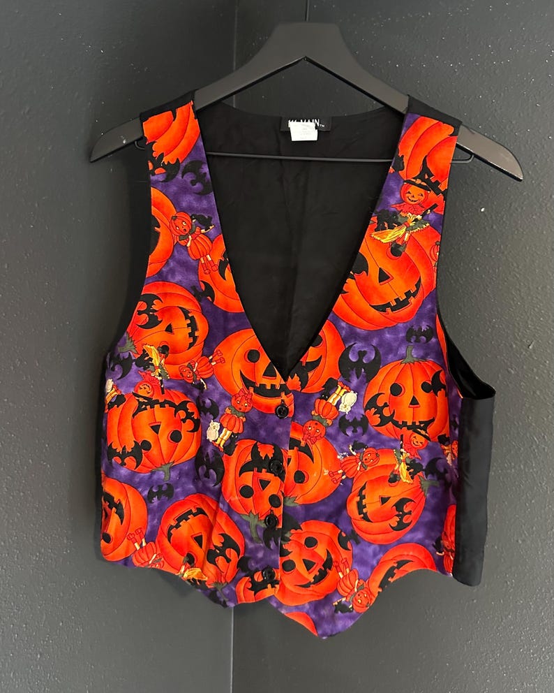 May include: A Halloween-themed vest with a black back and a vibrant front featuring orange jack-o'-lanterns on a purple background. The vest has a V-neck and is buttoned up, perfect for a festive outfit.