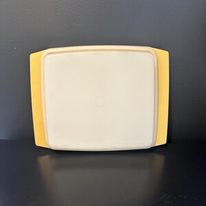 May include: A vintage yellow and off-white rectangular food storage container. The container has a square shape with rounded corners and a slightly raised edge. The lid is off-white, and the base is yellow.